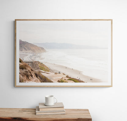 A slightly hazy, high-angle Landscape Beach Print captures a wide beachscape print where a light blue ocean waves meet a sandy shore dotted with numerous people enjoying the Summer Vibes. Gentle white waves roll towards the beach. Blue Lifeguard Towers line the coast in the far distance, of this framed landscape beach print. The overall lighting suggests a bright, overcast travel photography day on the San Diego coast. Photograph in minimalist mock up