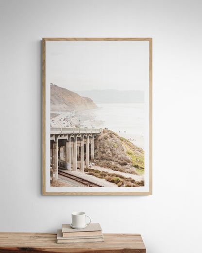 Captivating Torrey Pines San Diego coast scene, presented as vertical beach art. This piece of travel photography features a prominent bridge extending over a wide sandy beach with calm ocean waves. A coastal road winds along the base of rugged cliffs, with a lone coastal palm tree adding a touch of tropical essence to this Southern California seascape print. Photograph in minimalist mock up