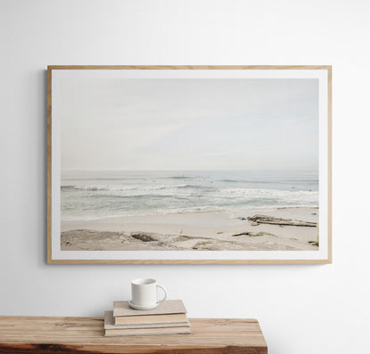 Fine art of horizontal landscape print by Southern California photographer Kim Dybczak of Salt Creek Prints, featuring a bright image captures a pale sandy beach giving way to a vast, light gray ocean with gentle waves. Several surfers are scattered across the water, some sitting on their boards and others riding small waves. The sky is a uniform, pale white, creating a soft, overcast atmosphere, located in Windansea Beach in La Jolla, California. Photograph in framed minimalist mock up
