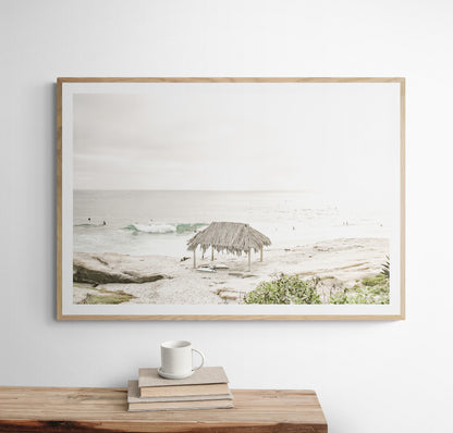 Fine art of WindanSea Beach, La Jolla California horizontal landscape print by Southern California photographer Kim Dybczak of Salt Creek Prints, featuring a bright, slightly desaturated image shows Windansea Beach in La Jolla, California. A thatched beach hut stands on the sandy shore in the left foreground. Further down the beach, a surfer carrying a teal surfboard walks towards the ocean where several other surfers are visible in the water. Photograph in minimalist framed mock up
