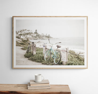 Fine art of horizontal landscape print by Southern California photographer Kim Dybczak of Salt Creek Prints, the image shows a light blue pastel beach cruiser leaning against a wooden post and chain fence. Another bicycle is visible behind it. The beach is in the background with people and a beach hut, located in Windansea Beach in La Jolla, California. Photograph in framed minimalist mock up