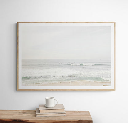 Fine art of horizontal landscape print by Southern California photographer Kim Dybczak of Salt Creek Prints, featuring a bright image captures a pale sandy beach giving way to a vast, light gray ocean with gentle waves. Several surfers are scattered across the water, some sitting on their boards and others riding small waves. The sky is a uniform, pale white, creating a soft, overcast atmosphere, located in Windansea Beach in La Jolla, California. Photograph in framed minimalist mock up