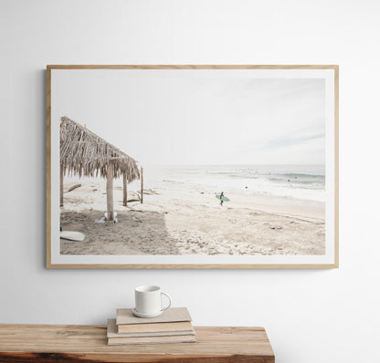 Fine art of WindanSea Beach, La Jolla California horizontal landscape print by Southern California photographer Kim Dybczak of Salt Creek Prints, featuring a bright, slightly desaturated image shows Windansea Beach in La Jolla, California. A thatched beach hut stands on the sandy shore in the left foreground. Further down the beach, a surfer carrying a teal surfboard walks towards the ocean where several other surfers are visible in the water. Photograph in minimalist framed mock up