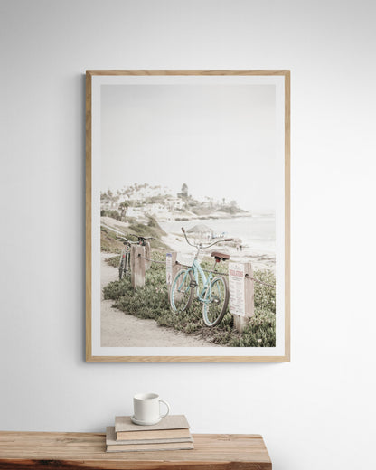 Fine art of horizontal landscape print by Southern California photographer Kim Dybczak of Salt Creek Prints, the image shows a light blue pastel beach cruiser leaning against a wooden post and chain fence. Another bicycle is visible behind it. The beach is in the background with people and a beach hut, located in Windansea Beach in La Jolla, California. Photograph in framed minimalist mock up