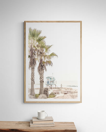 Dreamy coastal palm tree print featuring two tall palm trees with shaggy trunks in the foreground and a pastel blue lifeguard tower centered in the distance. This vertical tropical print is captured at Seaside Beach on the Southern California coast, is bathed in bright, hazy sunlight, creating a pastel beach art effect. Evokes serene summer vibes, perfect for beach house decor. Photograph in minimalist mock up.