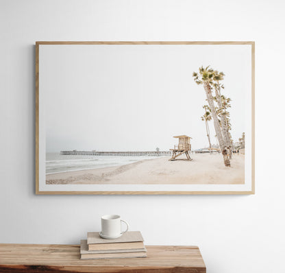 A wide landscape coastal photography view of San Clemente State Beach in Southern California, featuring a row of tall palm trees, a lifeguard tower, and people enjoying the calm beach. This image evokes Summer Vibes Wall Art and tropical beach print aesthetics. Photograph in minimalist mock up.
