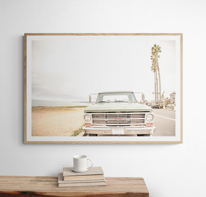 Nostalgic Southern California beach art featuring a classic light green Ford F-Series pickup truck parked by the San Diego coast. This horizontal surf print captures the vintage vehicle with a backdrop of iconic tall palm trees, a hazy beach, and the ocean, embodying surf vibes and perfect for coastal farmhouse decor. Photograph in minimalist framed mock up.