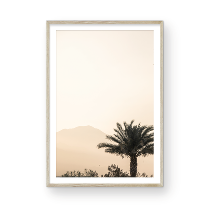 Vertical coastal print of a single palm tree silhouetted against a hazy sky with a distant mountain range, creating a calm beach print feel. Photograph in minimalist mock up.