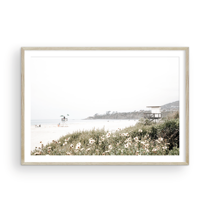 Landscape coastal photography of Salt Creek Beach Park, featuring a lifeguard tower and white flowers in the foreground, with distant cliffs. This calm beach print offers a serene coastal vibe. Photograph in minimalist mock up.