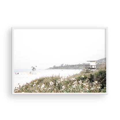 Landscape coastal photography of Salt Creek Beach Park, featuring a lifeguard tower and white flowers in the foreground, with distant cliffs. This calm beach print offers a serene coastal vibe. Photograph in minimalist mock up.