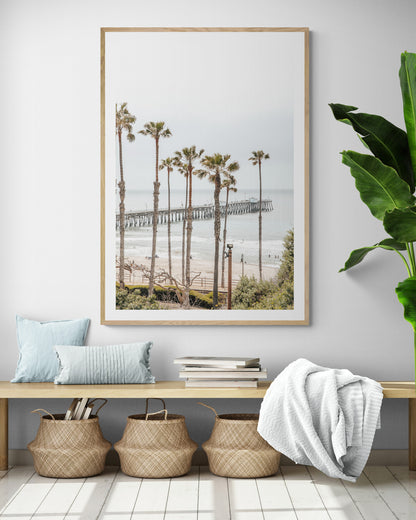A vertical landscape coastal photography shot of the San Clemente Pier from behind a lush line of tall palm trees. This beachscape print highlights the California coast and is ideal for large beach art or a beach theme wall decor, with subtle ocean waves. Photograph in minimalist mock up.