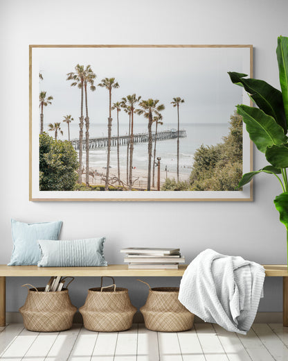 A horizontal landscape coastal photography shot of the San Clemente Pier from behind a lush line of tall palm trees. This beachscape print highlights the California coast and is ideal for large landscape beach art or a beach theme wall decor, with subtle ocean waves. Photograph in minimalist mock up.