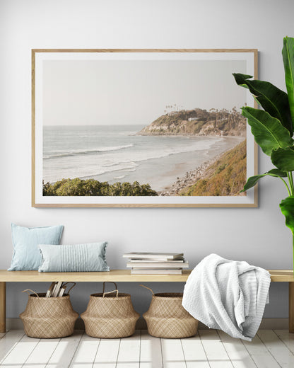 Fine art of Swamis Beach, San Diego California horizontal print of hazy high-angle landscape beach photography captures a light blue ocean meeting a sandy beach with many beach goers. To the right, tall tan and brown cliffs rise as iconic coastal scenery. 