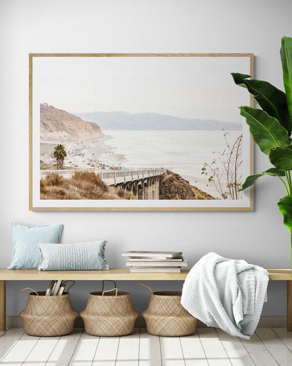 Captivating San Diego coast scene, presented as horizontal beach art. This piece of travel photography features a prominent bridge extending over a wide sandy beach with calm ocean waves. A coastal road winds along the base of rugged cliffs, with a lone coastal palm tree adding a touch of tropical essence to this Southern California seascape print. Photograph in minimalist mock up