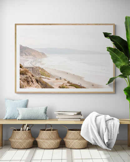  A slightly hazy, high-angle Landscape Beach Print captures a wide beachscape print where a light blue ocean waves meet a sandy shore dotted with numerous people enjoying the Summer Vibes. Gentle white waves roll towards the beach. Blue Lifeguard Towers line the coast in the far distance, of this framed landscape beach print. The overall lighting suggests a bright, overcast travel photography day on the San Diego coast. Photograph in minimalist mock up