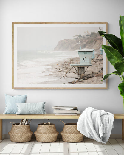 A serene piece of beach photography showcasing a light blue lifeguard tower, number 5, on the San Diego coast. This pastel beach art captures the iconic tower standing on wet sand with gentle ocean waves receding, under a hazy, overcast sky with distant cliffs. A perfect landscape lifeguard tower print for coastal theme wall art. Photograph in minimalist surf art mock up.