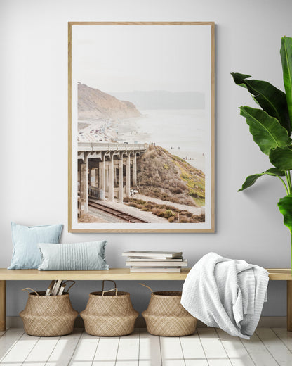 Captivating Torrey Pines San Diego coast scene, presented as vertical beach art. This piece of travel photography features a prominent bridge extending over a wide sandy beach with calm ocean waves. A coastal road winds along the base of rugged cliffs, with a lone coastal palm tree adding a touch of tropical essence to this Southern California seascape print. Photograph in minimalist entry way mock up