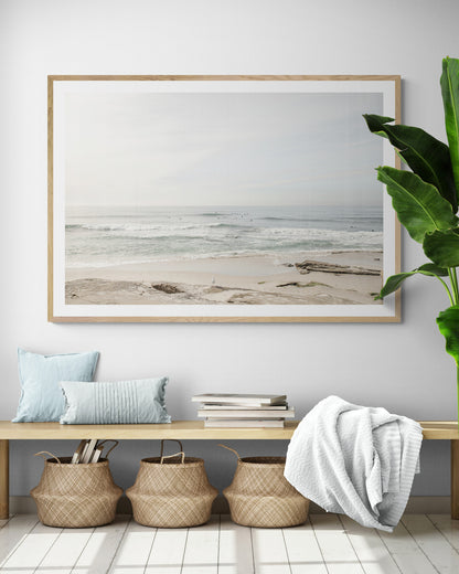Fine art of horizontal landscape print by Southern California photographer Kim Dybczak of Salt Creek Prints, featuring a bright image captures a pale sandy beach giving way to a vast, light gray ocean with gentle waves. Several surfers are scattered across the water, some sitting on their boards and others riding small waves. The sky is a uniform, pale white, creating a soft, overcast atmosphere, located in Windansea Beach in La Jolla, California. Photograph in framed minimalist mock up