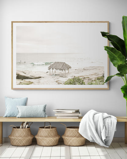 Fine art of WindanSea Beach, La Jolla California horizontal landscape print by Southern California photographer Kim Dybczak of Salt Creek Prints, featuring a bright, slightly desaturated image shows Windansea Beach in La Jolla, California. A thatched beach hut stands on the sandy shore in the left foreground. Further down the beach, a surfer carrying a teal surfboard walks towards the ocean where several other surfers are visible in the water. Photograph in minimalist framed entry way mock up
