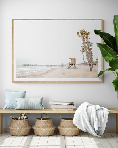 A wide landscape coastal photography view of San Clemente State Beach in Southern California, featuring a row of tall palm trees, a lifeguard tower, and people enjoying the calm beach. This image evokes Summer Vibes Wall Art and tropical beach print aesthetics. Photograph in minimalist mock up.