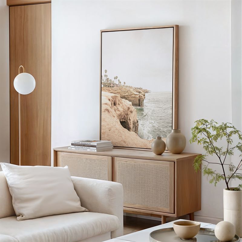 Modern living room with a wooden cabinet, white sofa, and framed artwork of a California coastal scene.