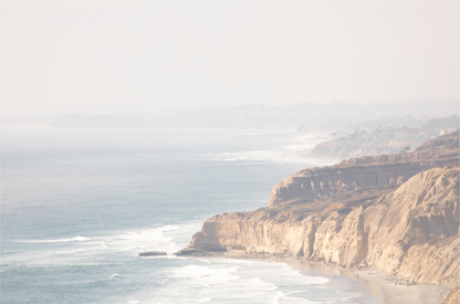 Fine art of Torrey Pines, San Diego California horizontal print of hazy high-angle landscape beach photography captures a light blue ocean meeting a sandy beach with many beach goers. To the right, tall tan and brown cliffs rise as iconic coastal scenery. 