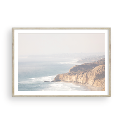 Fine art of Torrey Pines, San Diego California horizontal print of hazy high-angle landscape beach photography captures a light blue ocean meeting a sandy beach with many beach goers. To the right, tall tan and brown cliffs rise as iconic coastal scenery. Photograph in minimalist framed mock up.