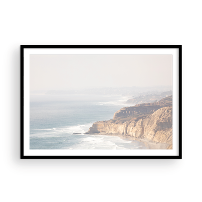 Fine art of Torrey Pines, San Diego California horizontal print of hazy high-angle landscape beach photography captures a light blue ocean meeting a sandy beach with many beach goers. To the right, tall tan and brown cliffs rise as iconic coastal scenery. Photograph in minimalist framed mock up.