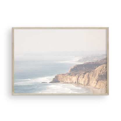 Fine art of Torrey Pines, San Diego California horizontal print of hazy high-angle landscape beach photography captures a light blue ocean meeting a sandy beach with many beach goers. To the right, tall tan and brown cliffs rise as iconic coastal scenery. Photograph in minimalist framed mock up.