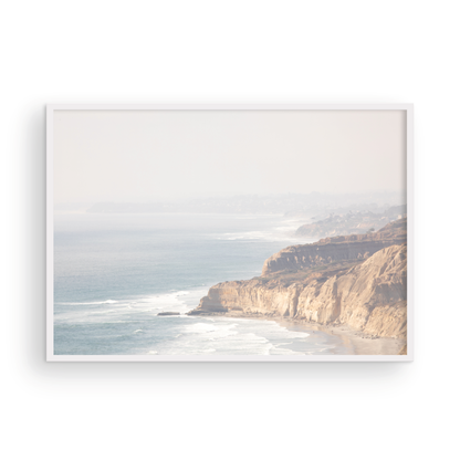 Fine art of Torrey Pines, San Diego California horizontal print of hazy high-angle landscape beach photography captures a light blue ocean meeting a sandy beach with many beach goers. To the right, tall tan and brown cliffs rise as iconic coastal scenery. Photograph in minimalist framed mock up.