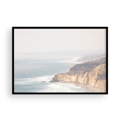 Fine art of Torrey Pines, San Diego California horizontal print of hazy high-angle landscape beach photography captures a light blue ocean meeting a sandy beach with many beach goers. To the right, tall tan and brown cliffs rise as iconic coastal scenery. Photograph in minimalist framed mock up.