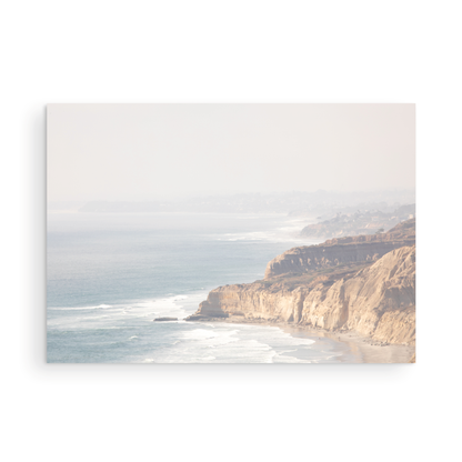 Fine art of Torrey Pines, San Diego California horizontal print of hazy high-angle landscape beach photography captures a light blue ocean meeting a sandy beach with many beach goers. To the right, tall tan and brown cliffs rise as iconic coastal scenery. Photograph in minimalist framed mock up.