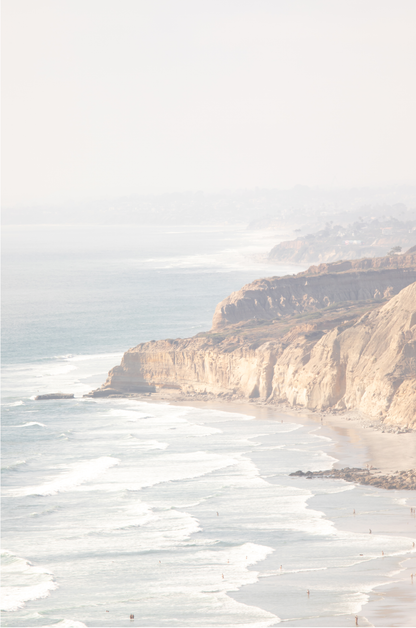 Fine art of Torrey Pines, San Diego California vertical print of hazy high-angle landscape beach photography captures a light blue ocean meeting a sandy beach with many beach goers. To the right, tall tan and brown cliffs rise as iconic coastal scenery.
