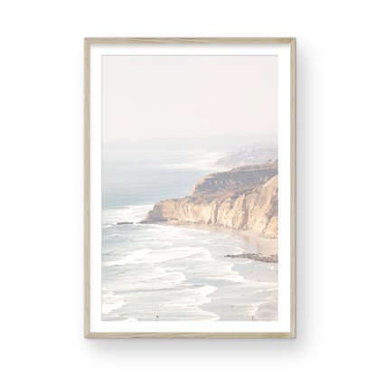 Fine art of Torrey Pines, San Diego California vertical print of hazy high-angle landscape beach photography captures a light blue ocean meeting a sandy beach with many beach goers. To the right, tall tan and brown cliffs rise as iconic coastal scenery. Photograph in minimalist framed mock up.