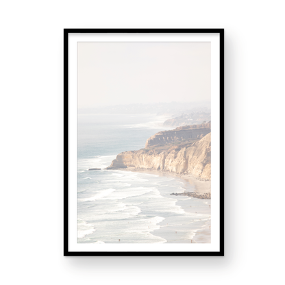 Fine art of Torrey Pines, San Diego California vertical print of hazy high-angle landscape beach photography captures a light blue ocean meeting a sandy beach with many beach goers. To the right, tall tan and brown cliffs rise as iconic coastal scenery. Photograph in minimalist framed mock up.