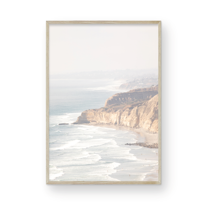 Fine art of Torrey Pines, San Diego California vertical print of hazy high-angle landscape beach photography captures a light blue ocean meeting a sandy beach with many beach goers. To the right, tall tan and brown cliffs rise as iconic coastal scenery. Photograph in minimalist framed mock up.