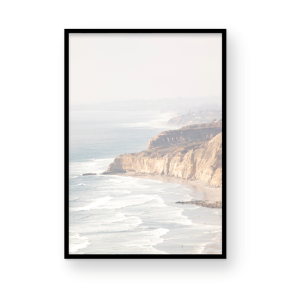 Fine art of Torrey Pines, San Diego California vertical print of hazy high-angle landscape beach photography captures a light blue ocean meeting a sandy beach with many beach goers. To the right, tall tan and brown cliffs rise as iconic coastal scenery. Photograph in minimalist framed mock up.