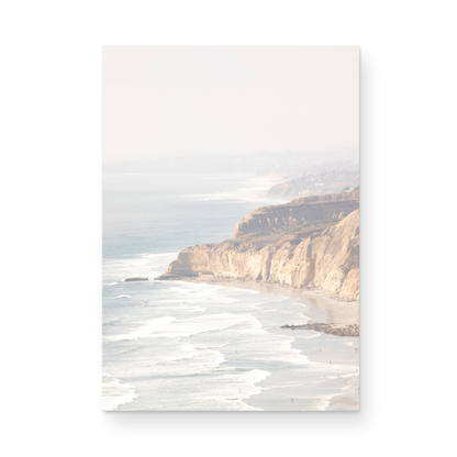 Fine art of Torrey Pines, San Diego California vertical print of hazy high-angle landscape beach photography captures a light blue ocean meeting a sandy beach with many beach goers. To the right, tall tan and brown cliffs rise as iconic coastal scenery. Photograph in minimalist framed mock up.