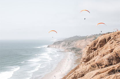 Fine art of Torrey Pines, San Diego California horizontal print of hazy high-angle landscape beach photography captures a light blue ocean meeting a sandy beach with many beach goers. To the right, tall tan and brown cliffs rise as iconic coastal scenery. Four white paragliders gliding in the pale pastel sky, a travel photography moment. The scene emphasizes the cliffs, beach, and the paragliders' presence in the expansive large beach art view. Photograph in minimalist mock up.