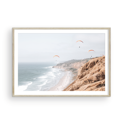 Fine art of Torrey Pines, San Diego California horizontal print of hazy high-angle landscape beach photography captures a light blue ocean meeting a sandy beach with many beach goers. To the right, tall tan and brown cliffs rise as iconic coastal scenery. Four white paragliders gliding in the pale pastel sky, a travel photography moment. The scene emphasizes the cliffs, beach, and the paragliders' presence in the expansive large beach art view. Photograph in minimalist wooden mock up.