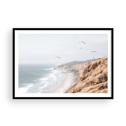 Fine art of Torrey Pines, San Diego California horizontal print of hazy high-angle landscape beach photography captures a light blue ocean meeting a sandy beach with many beach goers. To the right, tall tan and brown cliffs rise as iconic coastal scenery. Four white paragliders gliding in the pale pastel sky, a travel photography moment. The scene emphasizes the cliffs, beach, and the paragliders' presence in the expansive large beach art view. Photograph in minimalist mock up.