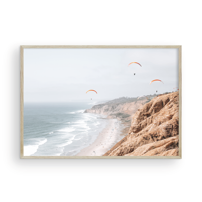 Fine art of Torrey Pines, San Diego California horizontal print of hazy high-angle landscape beach photography captures a light blue ocean meeting a sandy beach with many beach goers. To the right, tall tan and brown cliffs rise as iconic coastal scenery. Four white paragliders gliding in the pale pastel sky, a travel photography moment. The scene emphasizes the cliffs, beach, and the paragliders' presence in the expansive large beach art view. Photograph in minimalist mock up.