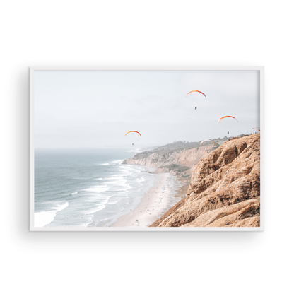 Fine art of Torrey Pines, San Diego California horizontal print of hazy high-angle landscape beach photography captures a light blue ocean meeting a sandy beach with many beach goers. To the right, tall tan and brown cliffs rise as iconic coastal scenery. Four white paragliders gliding in the pale pastel sky, a travel photography moment. The scene emphasizes the cliffs, beach, and the paragliders' presence in the expansive large beach art view. Photograph in minimalist mock up.