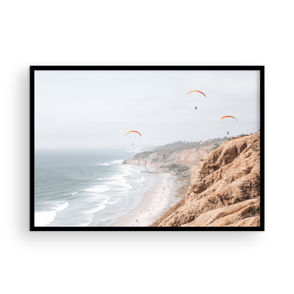 Fine art of Torrey Pines, San Diego California horizontal print of hazy high-angle landscape beach photography captures a light blue ocean meeting a sandy beach with many beach goers. To the right, tall tan and brown cliffs rise as iconic coastal scenery. Four white paragliders gliding in the pale pastel sky, a travel photography moment. The scene emphasizes the cliffs, beach, and the paragliders' presence in the expansive large beach art view. Photograph in minimalist mock up.