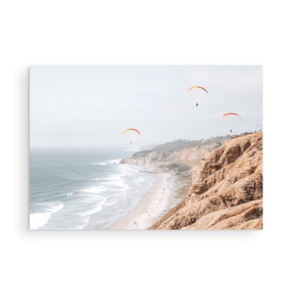 Fine art of Torrey Pines, San Diego California horizontal print of hazy high-angle landscape beach photography captures a light blue ocean meeting a sandy beach with many beach goers. To the right, tall tan and brown cliffs rise as iconic coastal scenery. Four white paragliders gliding in the pale pastel sky, a travel photography moment. The scene emphasizes the cliffs, beach, and the paragliders' presence in the expansive large beach art view. Photograph in minimalist mock up.