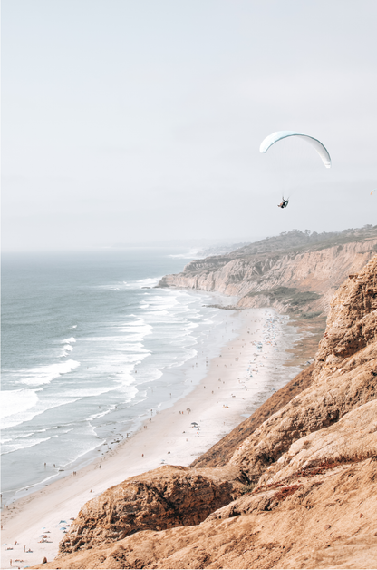 Fine art of Torrey Pines, San Diego California vertical print of hazy high-angle landscape beach photography captures a light blue ocean meeting a sandy beach with many beach goers. To the right, tall tan and brown cliffs rise as iconic coastal scenery. A white paraglider with a pilot flies in the upper right against a pale pastel sky, a travel photography moment. The scene emphasizes the cliffs, beach, and the paraglider's presence in the expansive large beach art view. Photograph in minimalist mock up