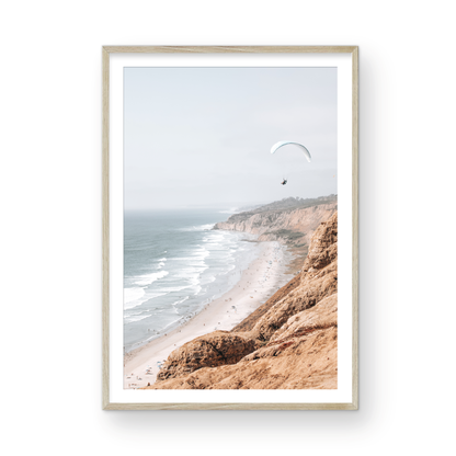 Fine art of Torrey Pines, San Diego California vertical print of hazy high-angle landscape beach photography captures a light blue ocean meeting a sandy beach with many beach goers. To the right, tall tan and brown cliffs rise as iconic coastal scenery. A white paraglider with a pilot flies in the upper right against a pale pastel sky, a travel photography moment. The scene emphasizes the cliffs, beach, and the paraglider's presence in the expansive large beach art view. Photograph in minimalist mock up