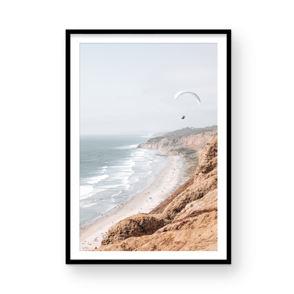 Fine art of Torrey Pines, San Diego California vertical print of hazy high-angle landscape beach photography captures a light blue ocean meeting a sandy beach with many beach goers. To the right, tall tan and brown cliffs rise as iconic coastal scenery. A white paraglider with a pilot flies in the upper right against a pale pastel sky, a travel photography moment. The scene emphasizes the cliffs, beach, and the paraglider's presence in the expansive large beach art view. Photograph in minimalist mock up