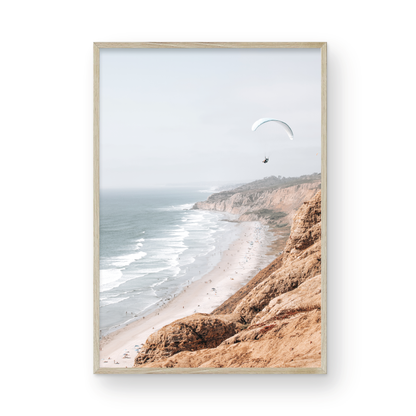 Fine art of Torrey Pines, San Diego California vertical print of hazy high-angle landscape beach photography captures a light blue ocean meeting a sandy beach with many beach goers. To the right, tall tan and brown cliffs rise as iconic coastal scenery. A white paraglider with a pilot flies in the upper right against a pale pastel sky, a travel photography moment. The scene emphasizes the cliffs, beach, and the paraglider's presence in the expansive large beach art view. Photograph in minimalist mock up