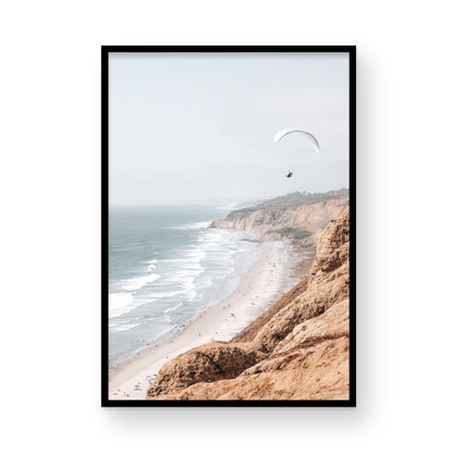 Fine art of Torrey Pines, San Diego California vertical print of hazy high-angle landscape beach photography captures a light blue ocean meeting a sandy beach with many beach goers. To the right, tall tan and brown cliffs rise as iconic coastal scenery. A white paraglider with a pilot flies in the upper right against a pale pastel sky, a travel photography moment. The scene emphasizes the cliffs, beach, and the paraglider's presence in the expansive large beach art view. Photograph in minimalist mock up